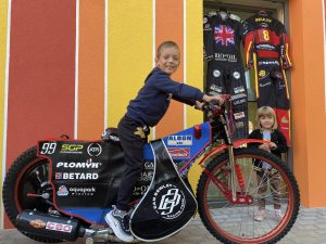 chłopiec na motorze . po prawej stronie dziewczynka. w tle widoczne stroje żużlowców.
