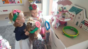 3 dziewczynki stoją przy szafce, patrzą na zabawkę i położone prezenty. 1 dziewczynka w sukience granatowej patrzy przez lupę. .