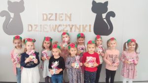 11 dziewczynek stoi obok siebie, trzymają w ręce prezenty. Na głowie mają opaski z czerwonym kwiatkiem.