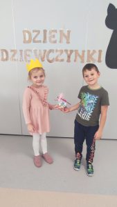 Stoi dziewczynka w różowej sukience, na głowie korona. Obok stoi chłopiec w zielonej bluzce ze wzorem. Wręcza prezent.