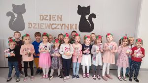 Dziewczynki i chłopcy stoją obok siebie. Dziewczynki na głowie mają opaskę z czerwonym kwiatkiem, w ręce trzymają prezent.