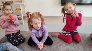 Na szarym dywanie siedzą 3 dziewczynki w bluzce czerwonej, fioletowej i różowej. W ręce ściskają kolorowe baloniki ,,Gniotki" są uśmiechnięte.