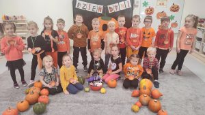 12 dzieci stoi, ubrani są w kolorach czarno-pomarańczowych, na szyi mają powieszone medale z dyni. Przed nimi siedzi 6 dzieci ubrane w kolorach czarno-żółto-pomarańczowych, na szyi mają powieszone medale z dyni. Przed nimi na szarym dywanie z dyni ułożone są dwie postacie z dyni.