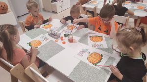 5 dzieci siedzi przy białym stole, na którym położone są zielone podkładki, na nich leżą kartki papieru z narysowaną dynią. Dzieci malują dynie farbami plakatowymi.
