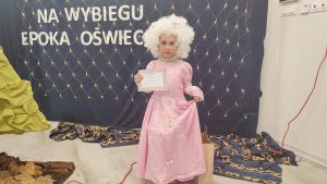 Dziewczynka z różowej sukni balowej i peruką. W ręce trzyma dyplom.