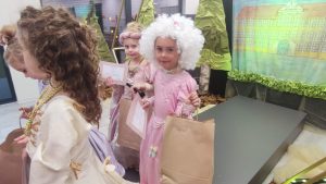 Dziewczynka z różowej sukni balowej i peruką. W ręce trzyma dyplom i torbę z prezentem.