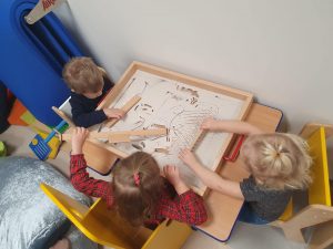 Dwie dziewczynki i chłopiec siedzący przy stoliku, bawiace sie piaskiem kinetycznym.