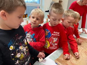 4 chłopców stoi obok siebie i rysują kredkami drzewo. Jeden chłopiec w czerwonej bluzie patrzy się na chłopca w granatowej bluzie.