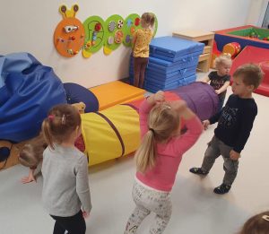 Dzieci uczestniczace w zabawach na sali rehabilitacyjnej. Dzewczynka przechodzi przez tunel, druga dziewczynka trzyma tunel, trzecia dziewczynka manipuluje ścianka sensoryczną. Dwie dziewczynki i chłopiec stoją obok tunelu.