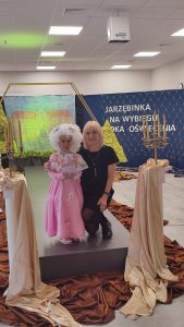 Dziewczynka z różowej sukni balowej z wachlarzem w ręce i peruką. Obok kuca nauczycielka.