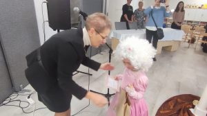 Pani Dyrektor wręcza dyplom dziewczynce w różowej sukni balowej i peruką.