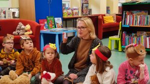 Pani w szarym swetrze klęczy i pokazuje dzieciom małą kartę do biblioteki. Naokoło niej siedzą dzieci ubrane na brązowo, na głowie mają czapki z misiami.