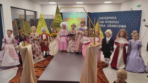 13 dzieci w strojach z epoki Oświecenia stoją na wybiegu i poza nim. W tyle za nimi zielone materiałowe choinki, szkic pałacu i granatowa tablica z napisem i lampkami. Przed dziećmi stoją duże świeczniki.