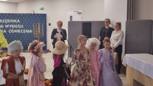 6 dzieci stoi ubrane w stroje z epoki Oświecenia. Za nimi stoi kobieta w czarnej marynarce i białej bluzce i przemawia do mikrofonu. Obok niej stoją 2 kobiety: jedna w czarnej sukience, druga w białej bluzce.