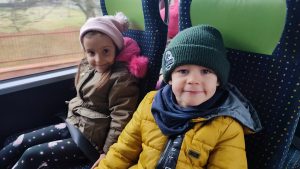 Chłopiec ubrany w kurtkę i czerwoną czapkę siedzi obok dziewczynki w niebieskiej kurtce i różowej czapce. Dzieci siedzą na fotelach w autobusie.