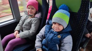 Chłopiec ubrany w kurtkę i szaro-zieloną czapkę siedzi obok dziewczynki w szarek kurtce i różowej czapce. Dzieci siedzą na fotelach w autobusie.