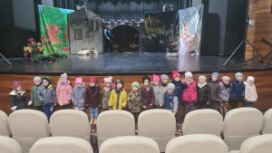 20 dzieci ubranych w kurtki i czapki stoją oparci o scenę teatralną, na której jest scenografia do bajki o Kocie w butach.