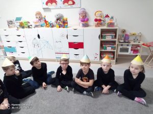 7 dzieci siedzi na szarym dywanie obok siebie. Ubrani są w czapki w kształcie kredki i czarne stroje.