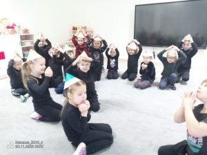 Dzieci w czarnych strojach, w czapkach w kształcie kredek siedzą na kolanach na szarym dywanie. Tworzą z rąk trójkąty nad głową.