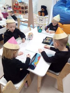 4 dzieci ubranych na czarno w czapkach w kształcie kredek, siedzą przy białym stole i rysują kredkami obrazek.