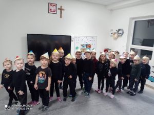 19 dzieci stoi w parach jeden za drugim. Dzieci ubrane są na czarno, na głowie mają czapki w kształcie kredek.