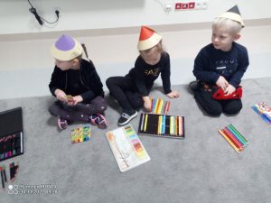 Troje dzieci ubranych na czarno siedzi na szarym dywanie. Na głowie mają czapki w kształcie kredek i układają kredki wg. wielkości.