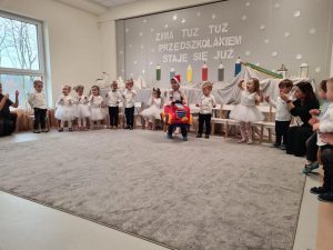 Dzieci ustawione w półkolu śpiewają piosenkę. Na środku dywanu, chłopiec w mikołajkowej czapce , jedzie samochodem