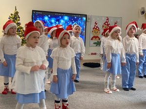 Dzieci ubrane w stroje biało-niebieskie stoją rozstawione na szarym dywanie i śpiewają piosenkę i kiwają rękami.