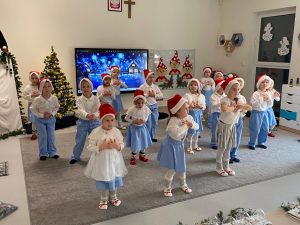 18 dzieci ubranych w stroje biało-niebieskie i czerwone czapki stoi w rozsypce na szarym dywanie i przyciągają do siebie splecione ręce. Dzieci tańczą.