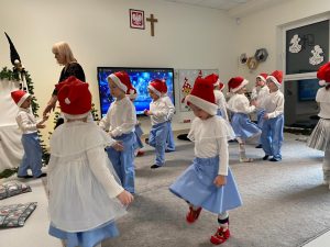 Dzieci stoją w parach na przeciw siebie i tupią nogami. Wszyscy ubrani są w czerwone czapki i strój biało-niebieski.