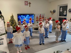 Dzieci ubrane w stroje biało-niebieskie stoją rozstawione na szarym dywanie i śpiewają piosenkę i kiwają rękami.