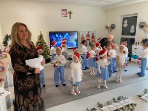 Na przodzie stoi pani dyrektor I. Hirsa, która trzyma w ręce białe dyplomy, ubrana jest w brązową sukienkę. Z tyłu na szarym dywanie stoją dzieci ubrane w czerwone czapki i strój biało-niebieski.