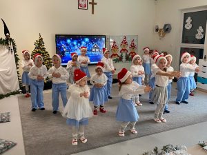 18 dzieci ubranych w stroje biało-niebieskie i czerwone czapki stoi w rozsypce na szarym dywanie i wyciągają przed siebie splecione ręce. Dzieci tańczą.