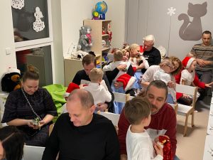 Dorosłe osoby siedzą na białych krzesłach i rozmawiają lub przytulają dziecko ubrane na biało -niebiesko w czerwonej czapce.