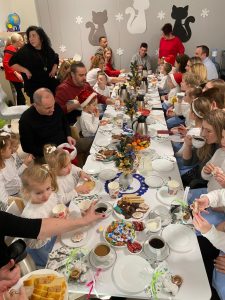 Przy białych stołach na których stoją słodkości bożonarodzeniowe siedzą dorosłe osoby wraz z dziećmi ubranymi na biało niebiesko.