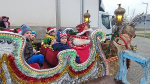 6 chłopców ubranych w kurtki siedzi w saniach Św. Mikołaja, które ciągnie brązowy renifer. Przed dziećmi siedzi Mikołaj ubrany w czerwony płaszcz i czapkę. Sanie ozdobione są świecidełkami.