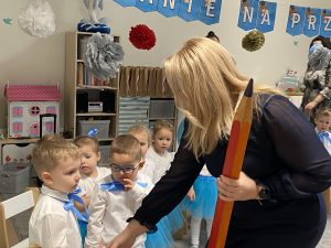 Pani Dyrektor trzymająca duży ołówek służący do pasowania, podajaca rękę przedszkolakowi. Po lewej stronie stoi pięcioro dzieci.