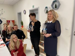 Pani Dyrektor Przedszkola nr 12 i Pani Dyrektor Wydziału Edukacji stojacy obok siebie i trzymający w rękach upominki. Z lewej strony przy stole siedzi pięcioro rodziców.