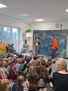 Duża grupa dzieci siedzi na dywanie i ogląda przedstawienie. Na środku stoi 2 aktorów przebranych za renifera i lisa.