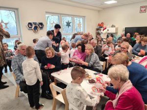 Zdjęcie przedstawia babcie i dziadków siedzących przy stołach i oglądających występy swoich wnuków, wszyscy są szczęśliwi i uśmiechnięci, sporo osób trzyma w ręku telefon i nagrywa.
