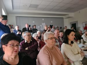 Na krzesłach siedzą starsi ludzie, kobiety i mężczyźni i patrzą przed siebie. Niektórzy z nich trzymają telefony i nagrywają.