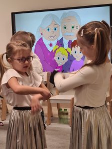 Dwie dziewczynki stoją na przeciw siebie, kręcą rękami tworząc młynek. Ubrane są w białe bluzki i srebrne spódnice.