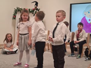 Dwóch chłopców ubranych w białe koszule i krawaty stoją obok siebie i mówią wiersz. Obok stoi dziewczynka ubrana na biało i srebrno. Jeden chłopiec odwrócony jest do dziewczynki.