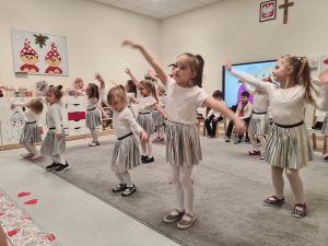 Dużo dziewczynek ubranych w białe bluzki i srebrne spódniczki stoją naprzemiennie na szarym dywanie i podnoszą jedną rękę do góry - tańczą.