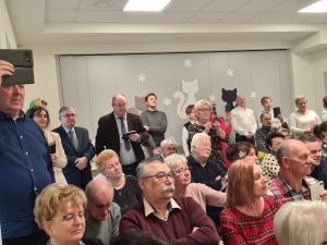 Na krzesłach siedzą starsi ludzie, kobiety i mężczyźni i patrzą przed siebie. Niektórzy z nich trzymają telefony i nagrywają.