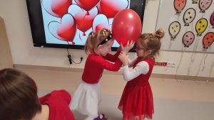 Chłopiec i dziewczynka ubrani na czerwono stoją plecami do siebie i pomiędzy trzymają czerwony balon. Tańczą do muzyki.