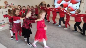 Dziewczynki ubrane na czerwono trzymają się za ramiona i stoją jedna za drugą. Chłopcy ubrani na czerwono stoją jeden za drugim i trzymają się za ramiona. Wszyscy poruszają się w rytm muzyki.