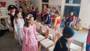 Na środku ustawione są krzesłya. Na około nich poruszają się dzieci w strojach karnawałowych w rytm muzyki.