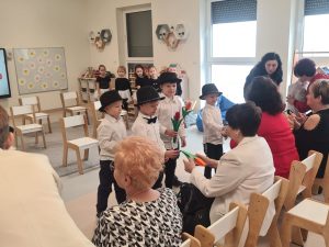 Chłopcy w białych koszulach i czarnych kapeluszach stoją przed zaproszonymi Paniami, i w rękach trzymają tulipany. Podchodzą i wręczają kwiaty.