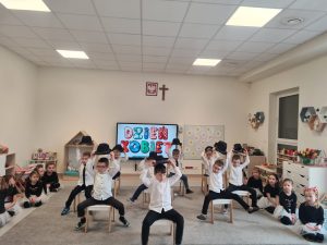 Chłopcy ubrani w białe koszule, siedzą na krzesłach. Na głowach mają czarne kapelusze, które podnoszą do góry do muzyki. Chłopcy pokazują taniec.